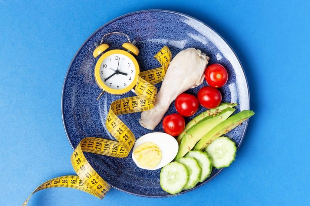 Plate with vegetables and yellow alarm clock on blue background